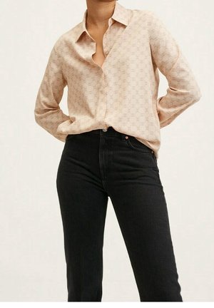 Kvinde iført en beige mønstret skjorte med knapper, stukket ned i højtaljede sorte jeans, med hænderne hvilende bag ryggen.