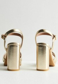 Sandali con tacco a blocco metallici dorati, con cinturino alla caviglia, texture liscia, punta aperta e un tacco rettangolare robusto. Dettaglio con fibbia regolabile.
