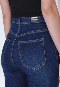 Jeans de mezclilla azul oscuro con cintura alta, costuras en contraste y un diseño clásico, que cuentan con una etiqueta negra de marca en la cintura trasera.