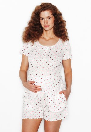 Donna incinta con capelli rossi ricci che indossa una maglietta bianca a maniche corte e pantaloncini coperti da piccoli cuori rossi, con una mano sulla pancia.