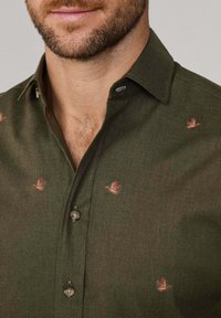 Man in een donkergroen shirt met rode en beige geborduurde eendenpatronen, gedeeltelijk ontknopt bij de kraag.