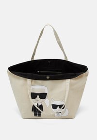 Beżowa torba płócienna z czarnym wnętrzem, z rysunkami kreskówkowymi przedstawiającymi mężczyznę i kota w okularach przeciwsłonecznych. Zawiera kieszeń zapinaną na zamek.