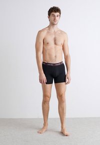 Čierne boxerky s logom na pásku, vyrobené z bavlneného materiálu, so priliehavým strihom a hladkou textúrou. Model stojí naboso na neutrálnom pozadí.