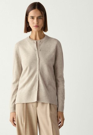 Frau mit kurzen braunen Haaren, die eine beige, geknöpfte Strickjacke und passende beigen Hosen trägt, steht vor einem schlichten hellen Hintergrund.