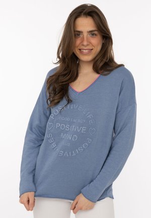 Frau mit langen braunen Haaren, die einen blauen Pullover mit dem Text "Positive Life, Good Energy, Positive Mind, Positive Vibes" und weißen Hosen trägt.
