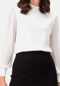 Femme portant un chemisier blanc avec des manches longues plissées transparentes et une jupe noire taille haute sur un fond uni.