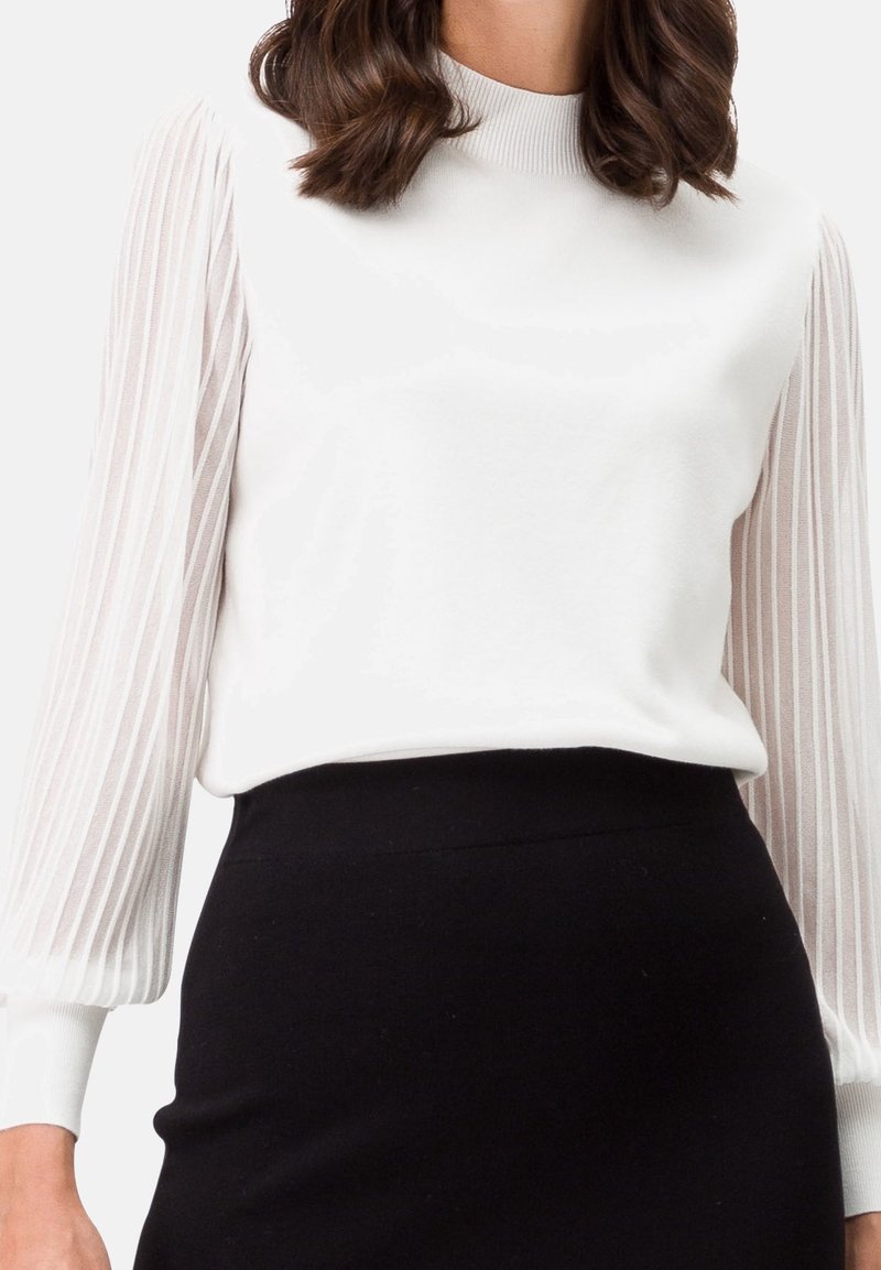 Femme portant un chemisier blanc avec des manches longues plissées transparentes et une jupe noire taille haute sur un fond uni.