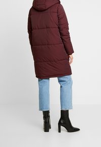 Cappotto puffer bordeaux con cappuccio, caratterizzato da un design trapuntato, maniche lunghe e una lunghezza fino al ginocchio, abbinato a jeans azzurri chiari e stivali neri alla caviglia.