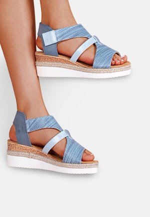 Pieds portant des sandales compensées tissées bleu clair avec des brides métalliques et des semelles intermédiaires à texture liège, semelles blanches en caoutchouc surélevées, sur un fond uni.