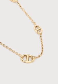 Κοντό χρυσό κολιέ με δύο οβάλ κρίκους, κάθε ένας με μια οριζόντια ράβδο στο κέντρο, σε απλό λευκό φόντο.