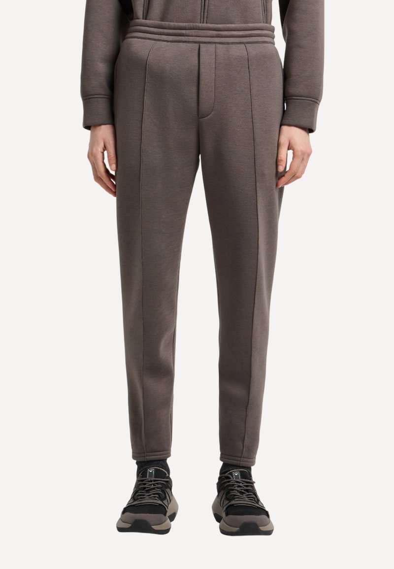 Pantaloni della tuta marroni con un design aderente, realizzati in materiale morbido, con pieghe frontali e gamba affusolata. Abbinati a scarpe da ginnastica nere.