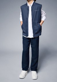 Námořnická vesty s dvěma předními kapsami přes bílou košili na knoflíky, v kombinaci s tmavými kalhotami a bílými teniskami; hladká struktura, ležérní design.
