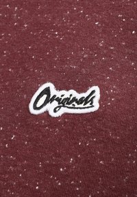 Bordová látka s bílými skvrnami a černobíle vyšívanou nášivkou se slovem "Originals" v kurzívním písmu.