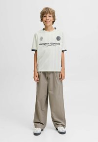 Chico de pie y sonriendo, vistiendo una camiseta gráfica blanca, pantalones beige holgados y zapatillas blancas sobre un fondo liso.