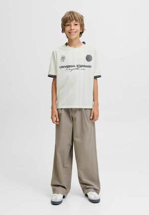 Garçon debout et souriant, portant un t-shirt graphique blanc, un pantalon beige ample et des baskets blanches sur un fond uni.