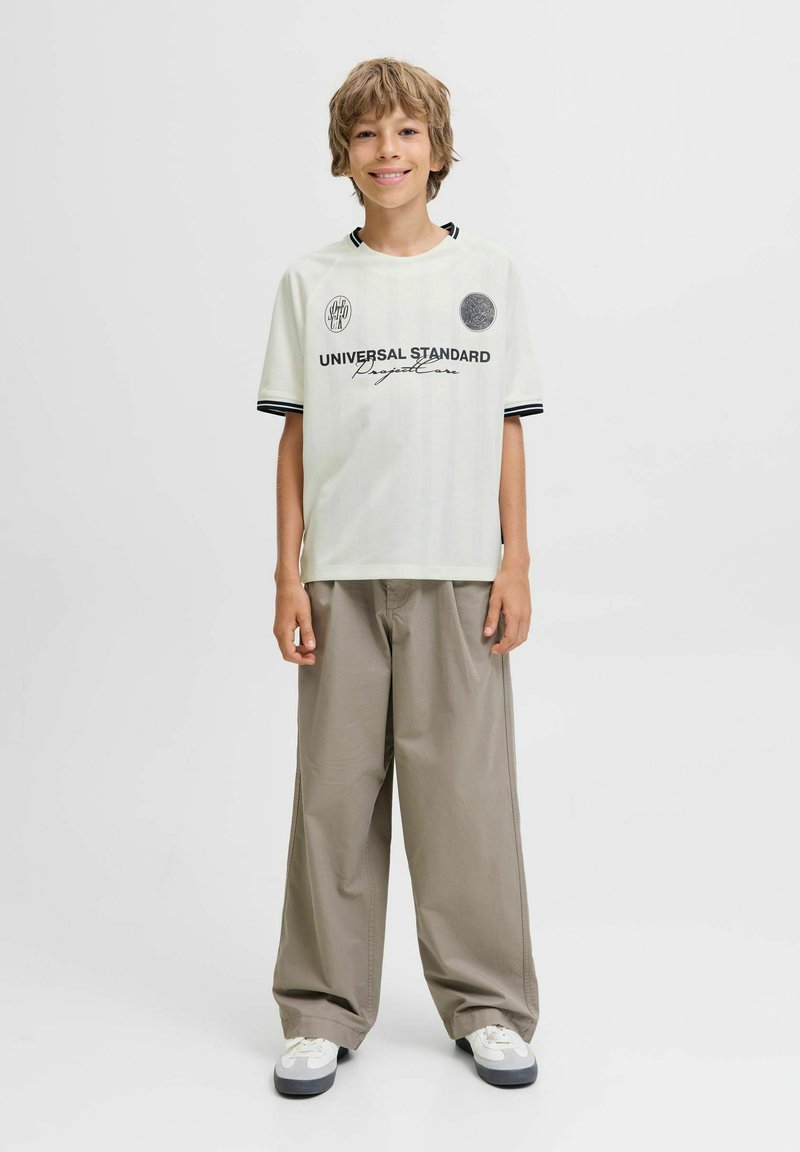 Chico de pie y sonriendo, vistiendo una camiseta gráfica blanca, pantalones beige holgados y zapatillas blancas sobre un fondo liso.
