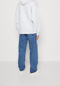 Person in einer hellgrauen Kapuzenjacke, lockeren blauen Jeans und weißen Turnschuhen, die vor einem einfarbigen weißen Hintergrund steht und sich wegdreht.