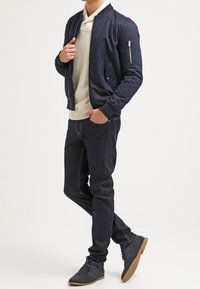 Granatowa kurtka bombowiec z prążkowanym kołnierzem, kieszenią na zamek, jasnoszary sweter, ciemne dżinsy i szare buty z jasnobrązowymi podeszwami. Ogólnie luźna stylizacja.