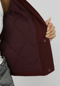 Doublure intérieure de la veste bordeaux avec un motif en vagues matelassées, dotée de boutons-pression en métal. Le tissu extérieur est lisse et résistant. Une main est visible tenant la veste.