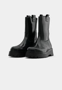 Schwarze ankle boots mit glänzender Oberfläche, ausgestattet mit einer kräftigen Sohle, einem seitlichen Reißverschluss und einem Schlaufen-Detail an der Ferse. Glatte Textur über das gesamte Paar.