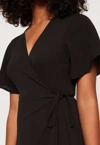 Černé obepínací šaty s krátkými volánovými rukávy, hlubokým V-výstřihem a zavázáním v oblasti pasu, vyrobené z jemné strukturované látky.