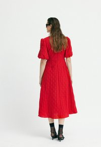 Rochie midi roșie cu o textură matlasată, mâneci bufante și talie sobră. Stilizată cu pantofi cu toc negri și ochelari de soare.