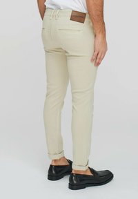 Pantaloni beige slim-fit in tessuto texturizzato, con tasche posteriori, risvolti e una patch in pelle marrone con il logo sulla vita.
