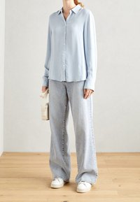 Camicia azzurra a maniche lunghe con bottoni e costine verticali, abbinata a jeans larghi in denim chiaro e sneakers bianche.