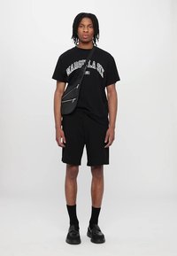 Fekete póló "MARGIELA SIX" felirattal, fekete sort, fekete bokazoknit és fekete cipőt visel. Ezenkívül egy fekete keresztpántos táska ezüst zipzárral.