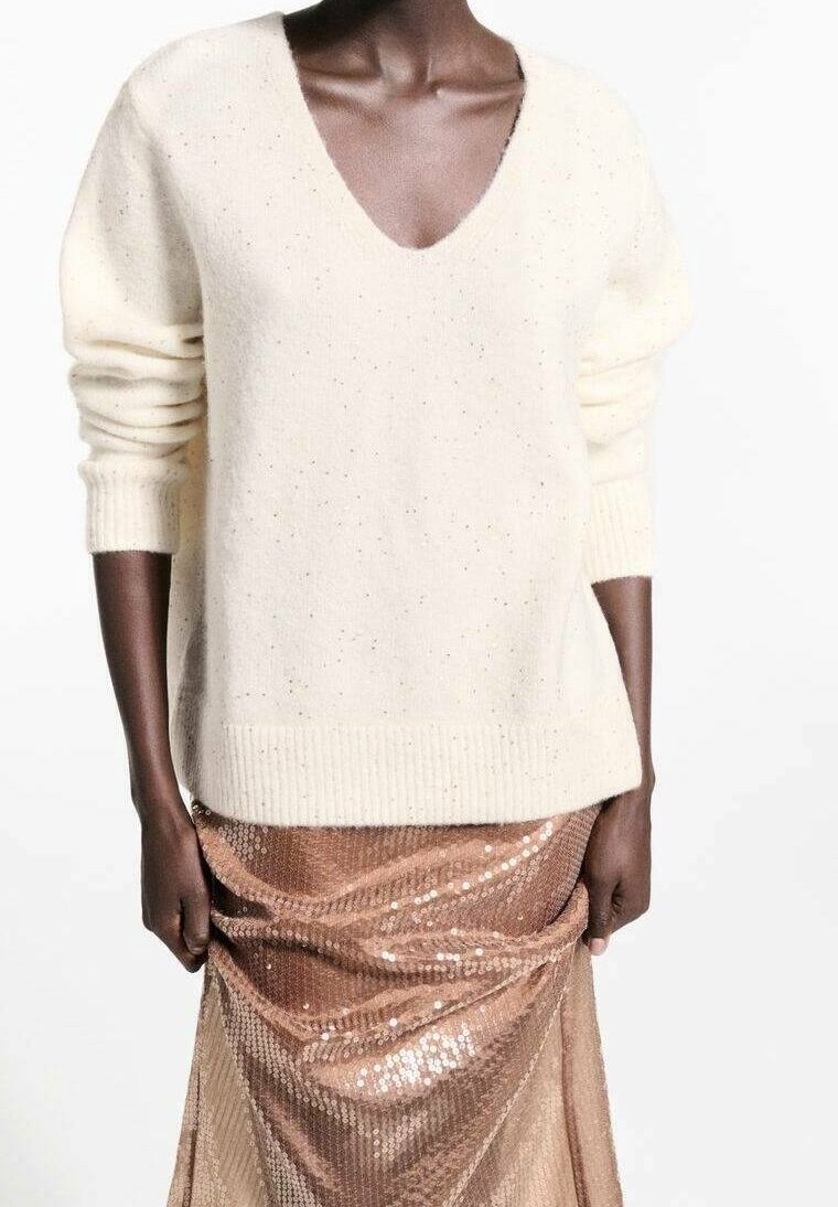 Personne à la peau foncée portant un pull crème à col en V avec poignets et ourlet côtelés, associé à une jupe brillante en sequins rose doré.