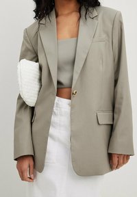 Light gray blazer made of smooth fabric, featuring a single button closure and two front pockets. Paired with a matching top and woven white clutch.