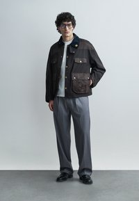 Moški z očali, temna srajca z gumbi in našitki, bela srajca, sive hlače in črni čevlji stoji pred enobarvnim ozadjem.