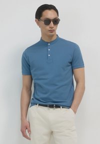 Polo blu a maniche corte con colletto alla cinese, dotato di tre bottoni. Indossato con pantaloni beige chiaro e occhiali da sole scuri.