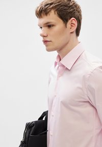 Ροζ ανοιχτό πουκάμισο με κουμπιά και κλασικό γιακά, κατασκευασμένο από λείο ύφασμα, με κουμπιά στο ίδιο χρώμα. Κρατιέται μία μαύρη τσάντα στο πλάι.