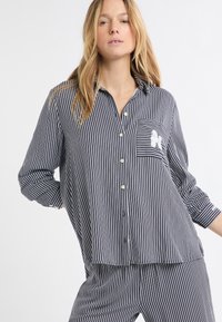 Koszula na guziki w biało-granatowe paski z kieszenią na piersi, ozdobiona niewielkim haftem, wykonana z lekkiego materiału o luźnym fasonie.