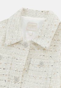 Weißes strukturiertes Jacket mit goldenen und silbernen Fäden, ausgestattet mit einem klassischen Kragen, Knopfverschlüssen und einem Etikett der Marke Guess.