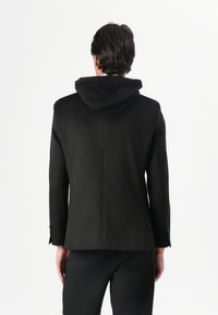 Schwarze Jacke mit Kapuze, strukturiertes Design, glatte Textur und ein Schlitz auf der Rückseite. Mit Knöpfen an den Bündchen.