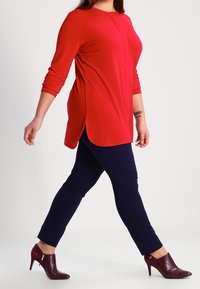 Röd långärmad topp med sidosprund, kombinerad med marinblå byxor och vinröda högklackade skor med dragkedjedetalj.