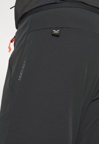 Svart utendørs bukser med elastisk midjebånd, rød snor, sidelomme og preget logo. Elastisk stoff med en glatt tekstur.