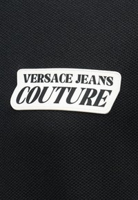 Patch blanche avec texte noir "VERSACE JEANS COUTURE" sur fond de tissu noir texturé.