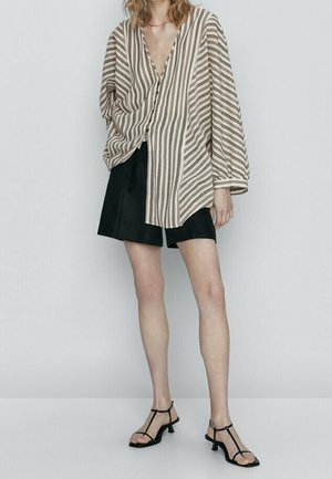 Femme portant une chemise oversize beige et marron à rayures boutonnée, un short noir et des sandales noires à petits talons, debout contre un mur uni.