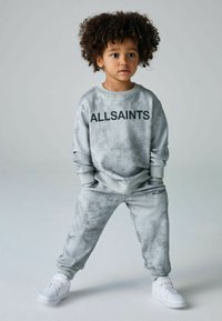 Pilkas medvilnės džemperis ir kelnės su švelnia tekstūra, dekoruoti juodu „ALLSAINTS“ logotipu, derinami su baltais sportbačiais.