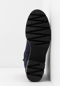 Vue de la semelle en caoutchouc noir avec des rainures en zigzag et des flèches, fixée à une chaussure bleu marine avec une fermeture éclair latérale visible.
