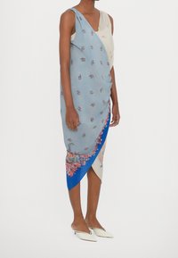 Robe bleue et crème à motifs floraux, ourlet asymétrique et design drapé. Associée à des chaussures blanches à bout pointu.