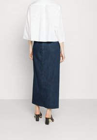 Bílá zkrácená košile spárovaná s tmavě modrou džínovou maxi sukni, která má hladkou strukturu. Outfit doplňují černé sandály na blokovém podpatku.