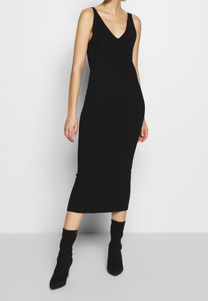 Kvinde iført en sort midi-kjole uden ærmer med V-formet halsudskæring, kombineret med sorte støvler til midten af læggen, stående mod en enkel, lys baggrund.