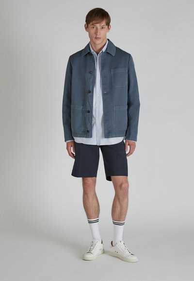 Blue casual jacket made of soft fabric, featuring a collar, front buttons, and two large pockets, worn over a striped shirt and shorts.