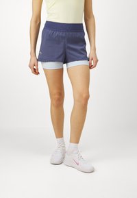 Ναυτικό μπλε σόρτς με φαρδύ ελαστικό, που περιλαμβάνει ανοιχτό μπλε εσωτερικά σορτς και φουσκωτές λεπτομέρειες κατά μήκος του στρινγκ. Φοριέται με λευκά αθλητικά παπούτσια.