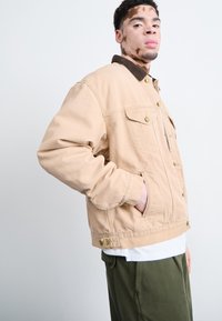 Chaqueta de denim beige con cuello de pana marrón, botones a presión, dos bolsillos frontales y un corte relajado. Llevada sobre una camisa blanca y pantalones verdes.