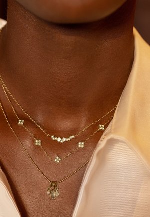 Drei zarte goldene Halsketten mit kleinen Edelstein-Anhängern, geschichtet auf einem dunkelhäutigen Hals, kombiniert mit einem cremefarbenen Hemd mit offenem Kragen.